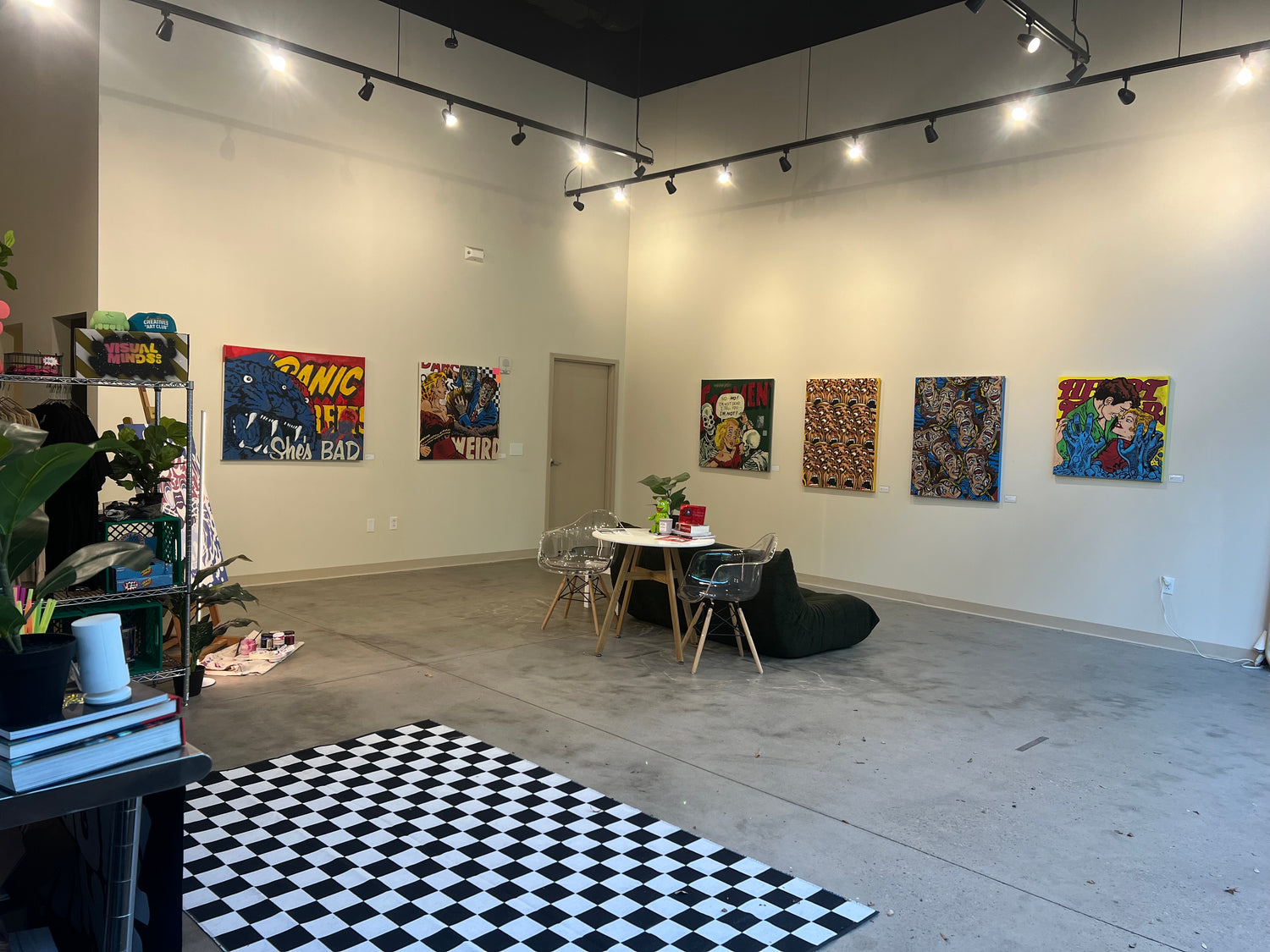 Art gallery with colorful paintings on white walls, a black and white checkered floor, and a small table with chairs.